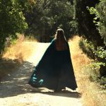 Jeune femme partant sur un chemin au milieu de la verdure, sa cape verte flotte au vent