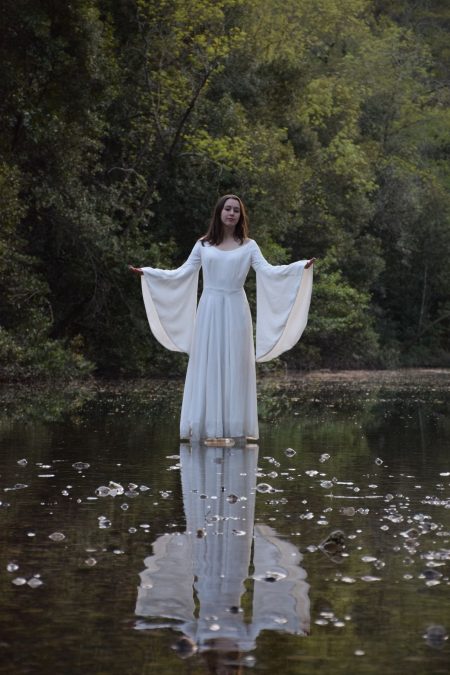 Jeune femme bras ouverts dans sa robe blanche féérique, ses longues manches évasées translucides dans la lumière du soir sur la rivière