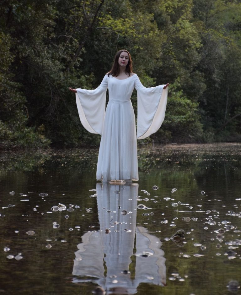Jeune femme bras ouverts dans sa robe blanche féérique, ses longues manches évasées translucides dans la lumière du soir sur la rivière
