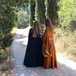 Deux jeunes filles en robe médiévale fantastique: l'une orange et l'autre bleue, de dos face au chemin bordé de cyprès
