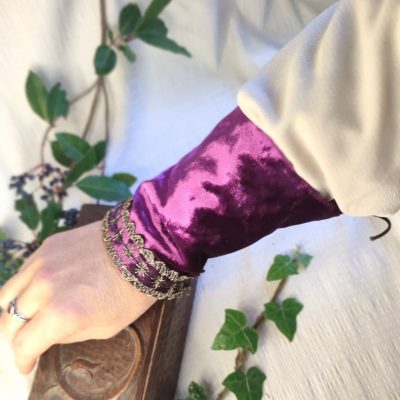 Bras d'une personne en gros plan portant un manchon de poignets fantasy de conte de fée en velours pourpre décorée d'un galon brodé or, posée sur un tissu crème à côté de petites branches avec des bais colorée