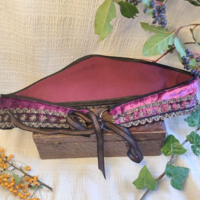 Ceinture fantasy de conte de fée en velours pourpre de dos ,décorée d'un galon brodé or, posée sur un tissu crème à côté de petites branches avec des bais colorées