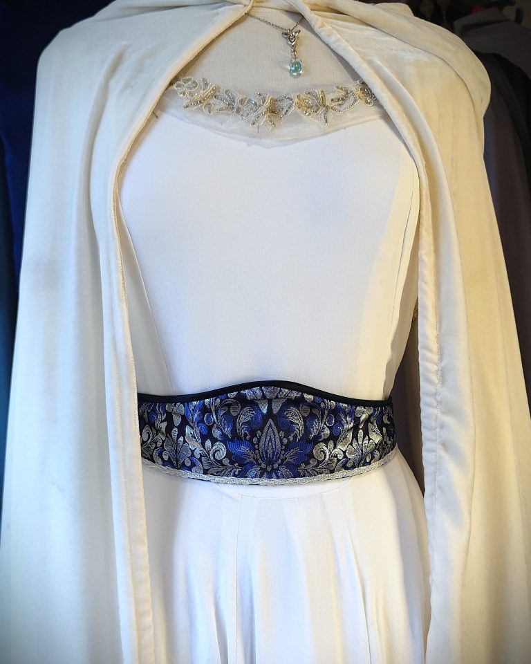 gros plan sur le buste d'un mannequin vêtu d'une robe blanche avec encoure brosée de perles et cape en velours et ceinture en brocard bleu argent